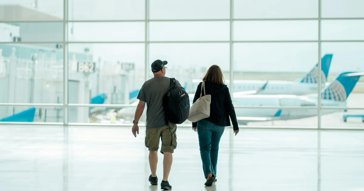 To reisende med bagasje som går gjennom en flyplassterminal med fly utenfor