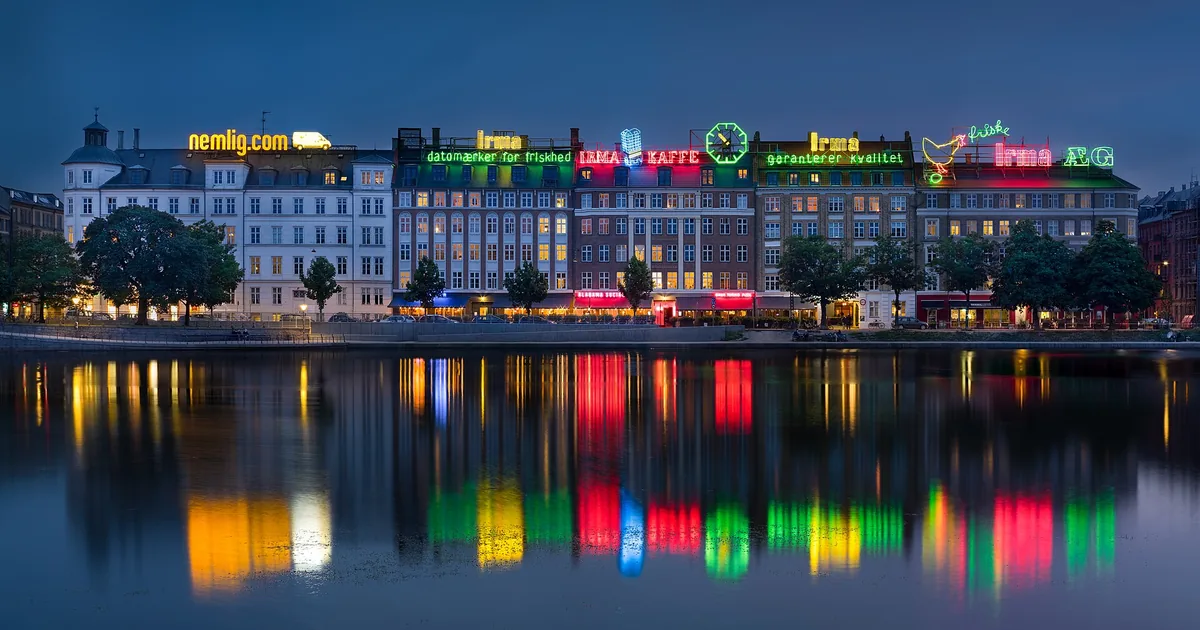 Opplyste reklameskilt som speiler seg i vannet foran en dansk bygningsrekke om kvelden